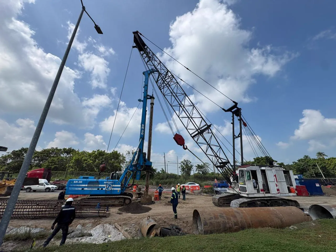 Equipo de perforación azul en un frente de obra vial con cielo abierto y personal de campo.